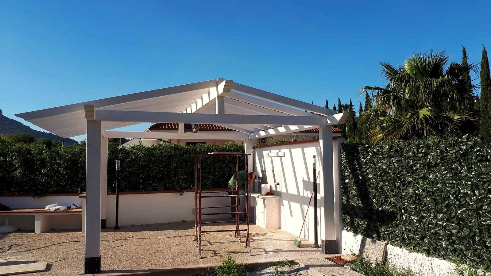 Pergola in legno bianco su un terrazzo con siepe e palme